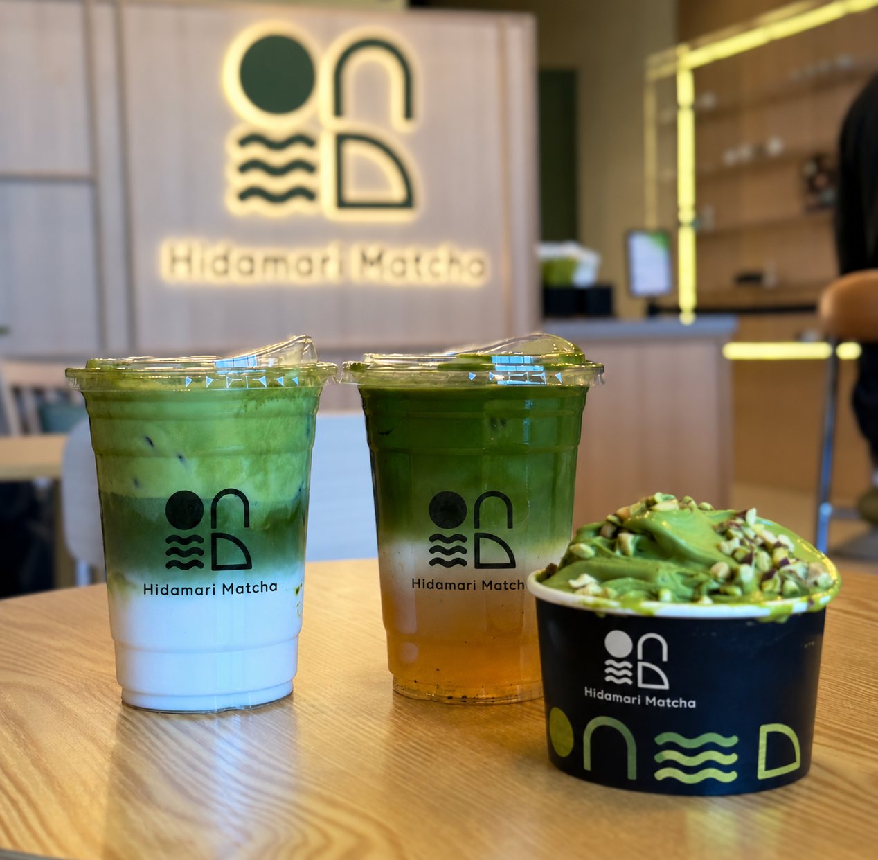 Two matcha drinks and a cup of pistachio gelato in front of the glowing Hidamari Matcha logo wall