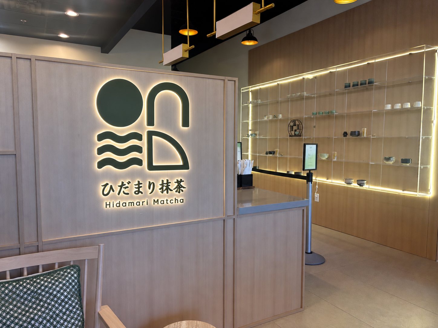 The backlit Hidamari Matcha logo on a pale oak wall beside a lit shelf of tea bowls